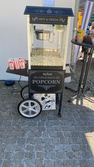 Popcornmaschine Kindergartenfest Popcornmaschine auf einem Wagen mit weißen Rädern und rotem Popcornbehälter daneben.
