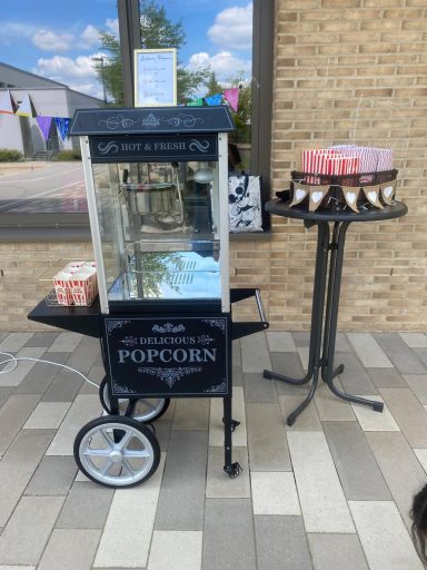 Popcornmaschine Sommerfest Popcornwagen mit Glaskabine und einem Tisch mit Popcorntüten in einem Außenbereich.